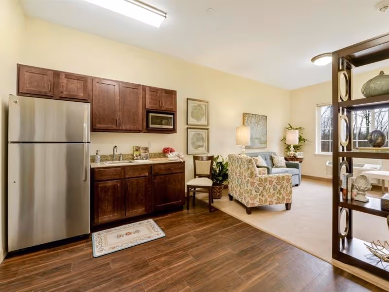 A cozy interior space featuring a small kitchen area with dark wooden cabinets, a stainless steel refrigerator, a microwave, and a sink. Adjacent to the kitchen is a living area with a floral patterned armchair, a blue sofa, a wooden chair, a floor lamp, and decorative plants. A shelving unit partially separates the kitchen and living area. The room has wood flooring in the kitchen section and carpet in the living area, with a window letting in natural light.