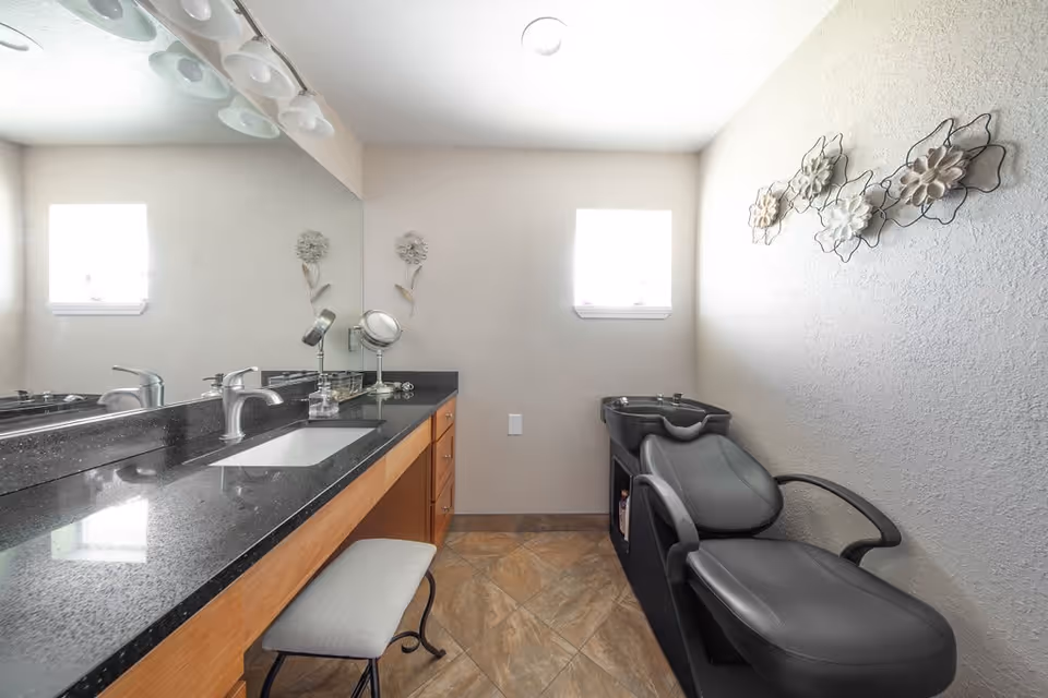 A clean and modern bathroom area featuring a long black granite countertop with a built-in sink, a large mirror above, and a cushioned stool underneath. To the right, there is a black salon-style chair with a built-in sink for hair washing. The walls are light-colored with decorative floral wall art, and there are two small windows letting in natural light.