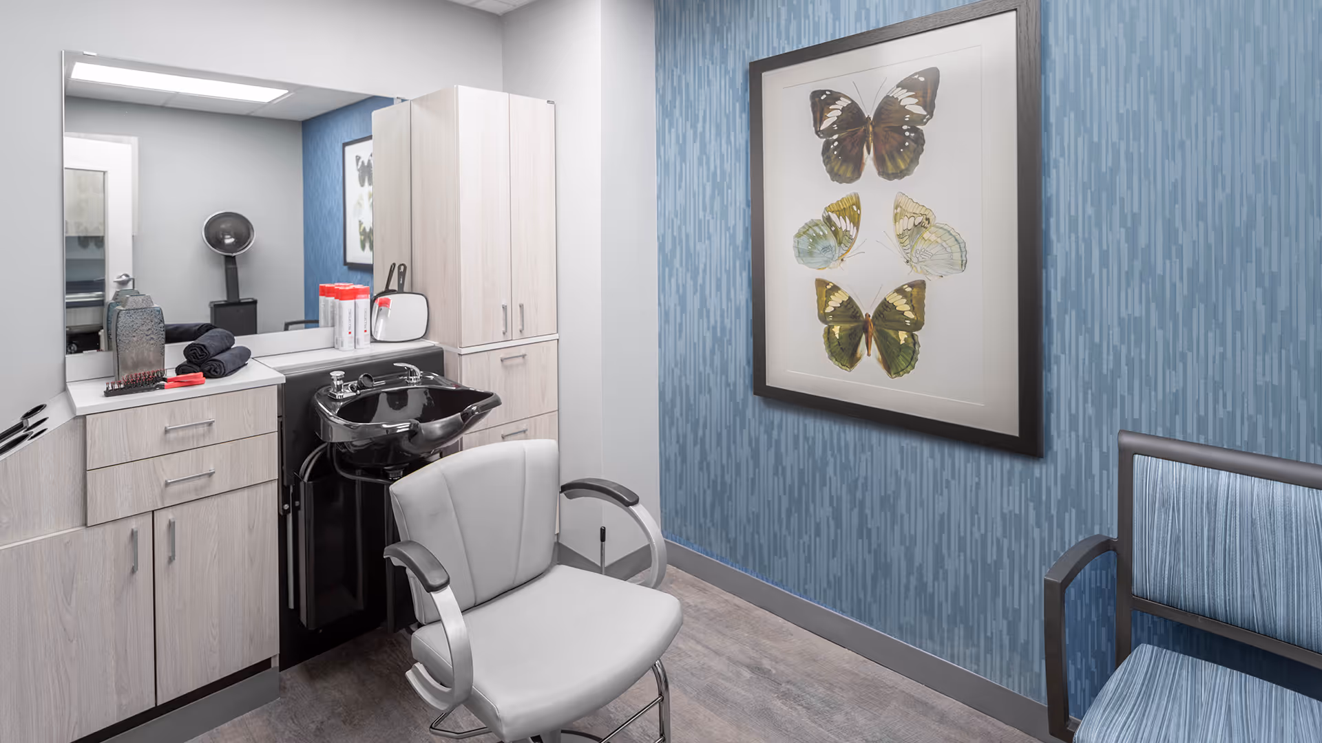 Interior of a small salon area with a gray salon chair in front of a black hair washing sink. Light wood cabinets and a large mirror are on the left wall. On the right wall, there is blue patterned wallpaper with a framed picture of butterflies. A blue upholstered chair is partially visible on the right side.
