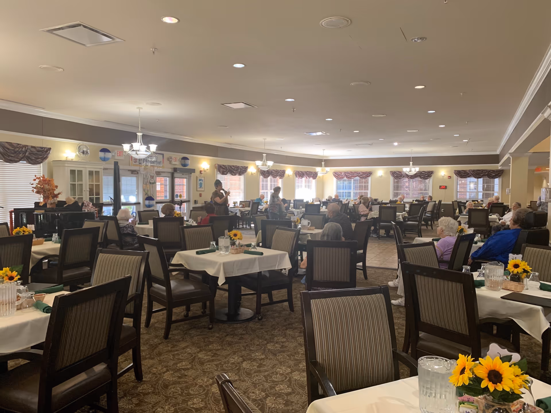 A spacious dining room in a senior living facility with multiple tables covered in white tablecloths, each decorated with a small vase of sunflowers. Several elderly residents are seated at the tables, some engaged in conversation. The room is well-lit with chandeliers and natural light from windows with valances. There is a piano in the corner and a few staff members are standing and interacting with the residents.