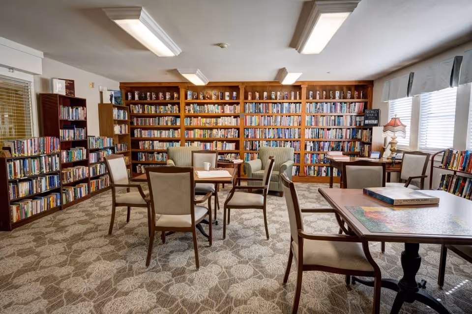 A cozy library room with multiple bookshelves filled with books along the walls. There are several tables with chairs arranged around them, and two armchairs near the back wall. The room is well-lit with ceiling lights and natural light coming through the windows on the right side. A table in the foreground has a puzzle and a book on it.