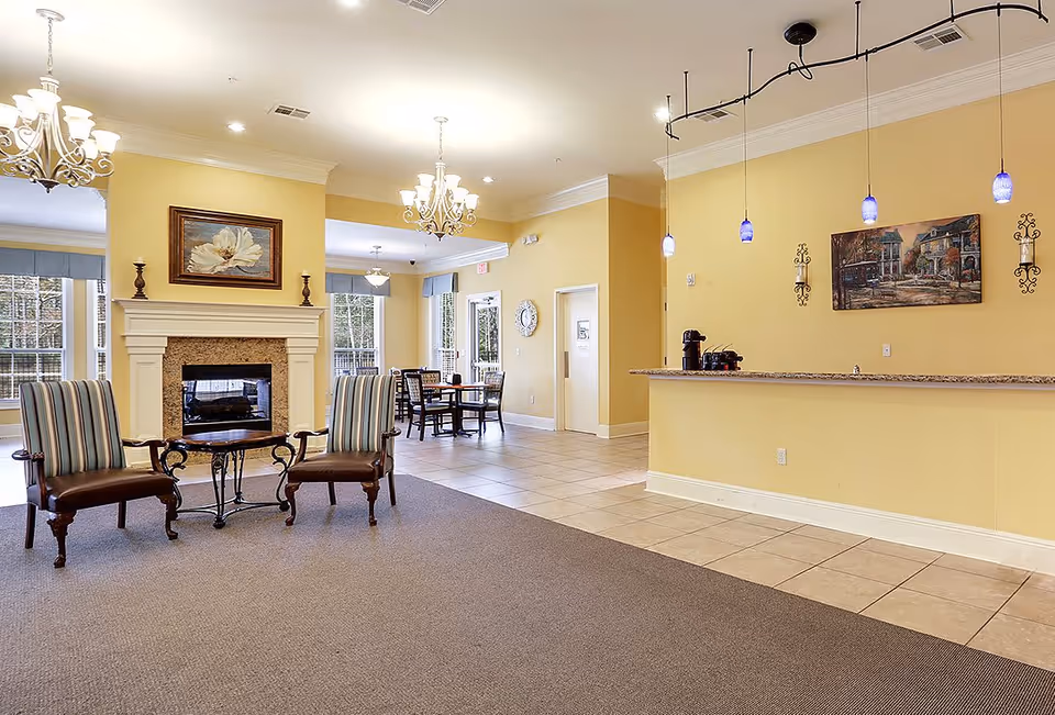 A spacious and well-lit common area in a senior living facility featuring two striped armchairs with wooden legs and arms, a small wooden table between them, a fireplace with a floral painting above it, a dining area with tables and chairs in the background, and a counter with hanging blue pendant lights and a coffee maker on it. The walls are painted yellow and the floor is a combination of carpet and tile.