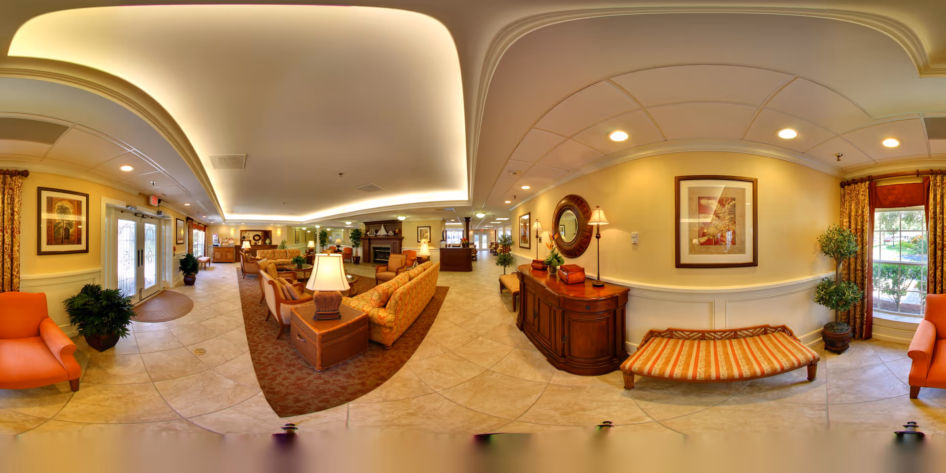 A spacious and warmly lit senior living facility common area with comfortable seating including sofas and armchairs arranged around a fireplace. The room features tiled floors, decorative plants, framed artwork on the walls, and large windows with curtains allowing natural light. There is a wooden sideboard with a round mirror and lamps, and the entrance door is visible on the left side.