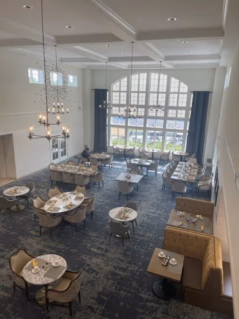 A spacious dining room with multiple round and rectangular tables set with plates, cups, and napkins. The room features high ceilings with chandeliers, large windows allowing natural light, and a carpeted floor. There are cushioned chairs around the tables and booth seating along one wall.