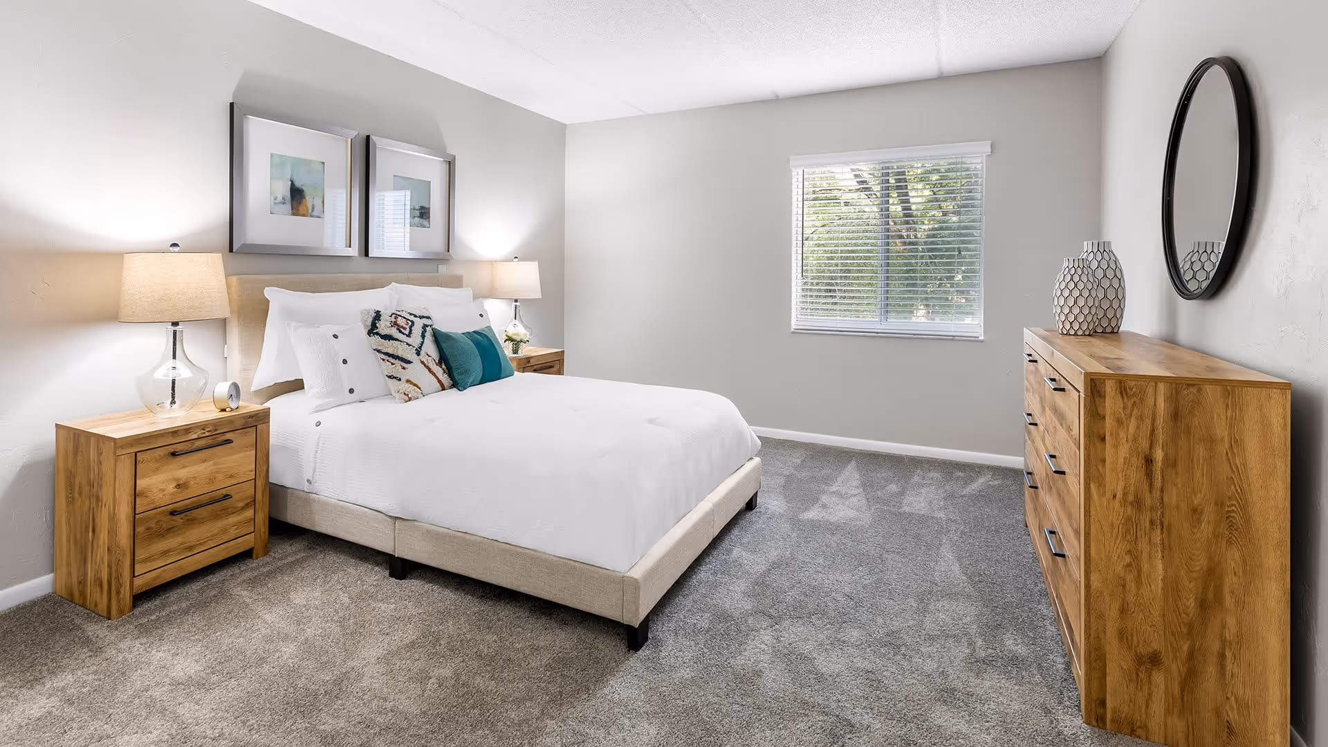 A bright and clean bedroom with a beige upholstered bed featuring white bedding and decorative pillows. There are two wooden nightstands with glass lamps on either side of the bed. Above the bed are two framed abstract artworks. A window with white blinds lets in natural light. On the right side of the room is a wooden dresser with a round mirror and two decorative vases on top. The floor is carpeted in gray.