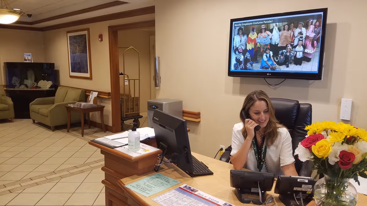 Reception area at Neville Center with a woman sitting at the front desk talking on the phone. The desk has a computer, phone, hand sanitizer, and a vase with colorful flowers. Behind her on the wall is a TV screen displaying a group photo titled 'Neville Employee Costume Parade!'. In the background, there are green armchairs, a side table, and an aquarium.