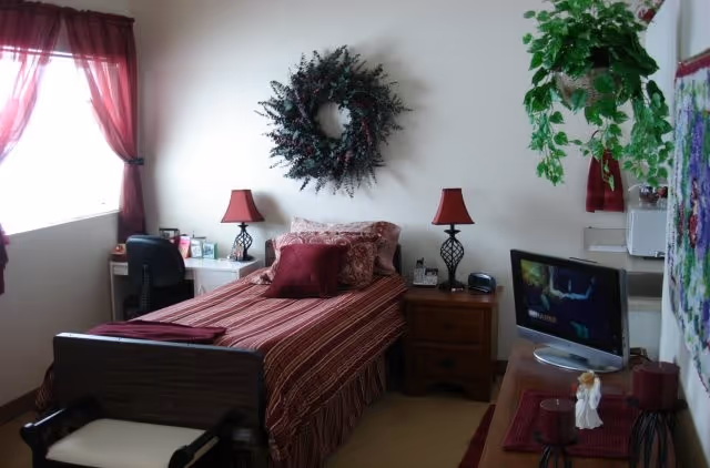 A cozy bedroom in an assisted living facility with a single bed covered in a red and beige striped bedspread and multiple pillows. There is a decorative wreath hanging on the wall above the bed. Two matching red lamps sit on bedside tables on either side of the bed. A desk with a black chair is positioned near a window with red curtains. A TV is placed on a dresser opposite the bed, and a green plant hangs from the ceiling near the window.