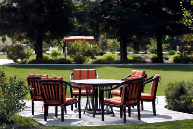 A circular patio in a grassy garden with a round table surrounded by six red-and-orange striped cushioned chairs.