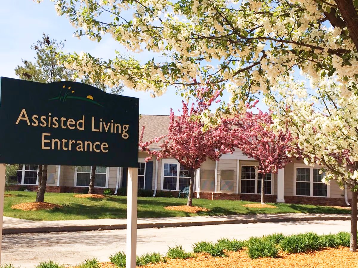 A green 'Assisted Living Entrance' sign in front of a single-story retirement community building with blooming pink and white trees.