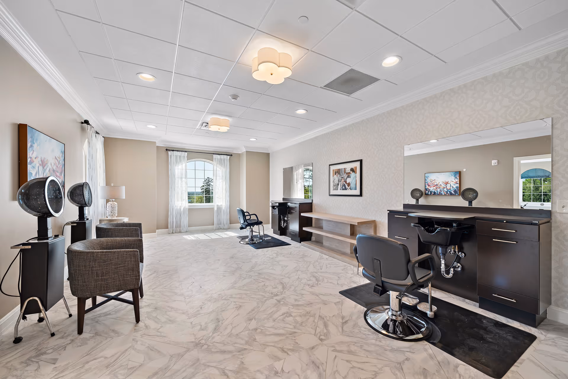 A bright and spacious hair salon area with two black salon chairs in front of large mirrors and wash basins. The room has light-colored marble flooring, beige walls, and a large window with sheer white curtains letting in natural light. Two hair dryers and two gray armchairs are positioned along one wall, with framed artwork hanging above. The ceiling has recessed lighting and decorative light fixtures.