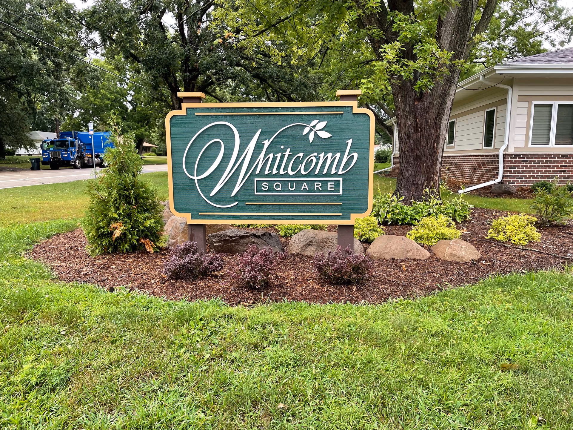 A green and beige sign reading 'Whitcomb Square' is displayed in a landscaped area with small bushes, rocks, and mulch. Behind the sign, there is a large tree and part of a building with beige siding and brick foundation. A street and a blue truck are visible in the background.