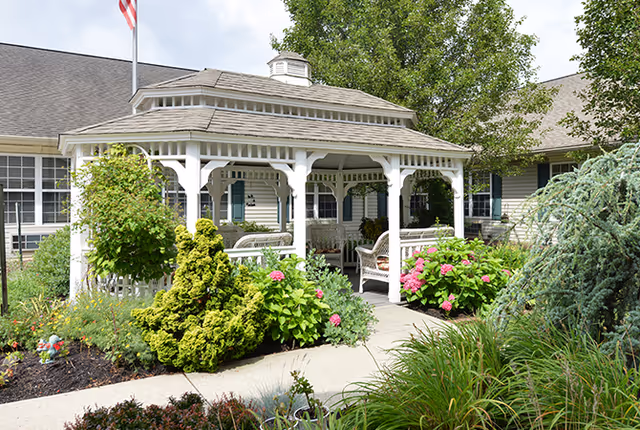 A white wooden gazebo surrounded by lush green bushes and colorful flowers in a garden area outside a building with beige siding and multiple windows. There are benches inside the gazebo and a concrete pathway leading to it. Trees and an American flag are visible in the background.