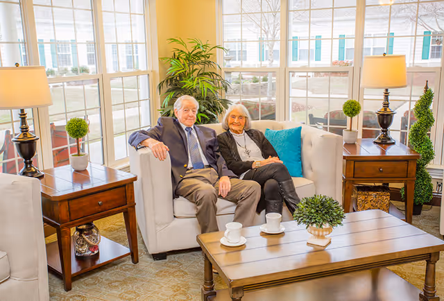 An elderly man and woman sitting together on a beige sofa in a bright living room with large windows. The room is decorated with wooden side tables, table lamps, small potted plants, and a wooden coffee table with two white cups and saucers. There is a large green plant behind the sofa and a blue cushion on the sofa.