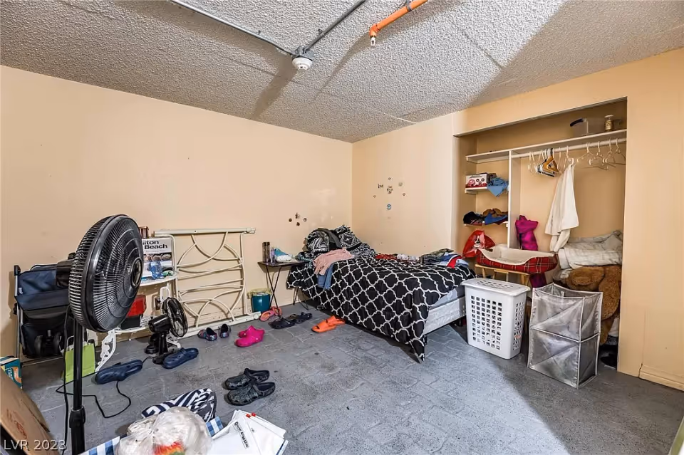 Cluttered bedroom with a single bed, open closet, laundry baskets, a standing fan and scattered shoes on the floor.