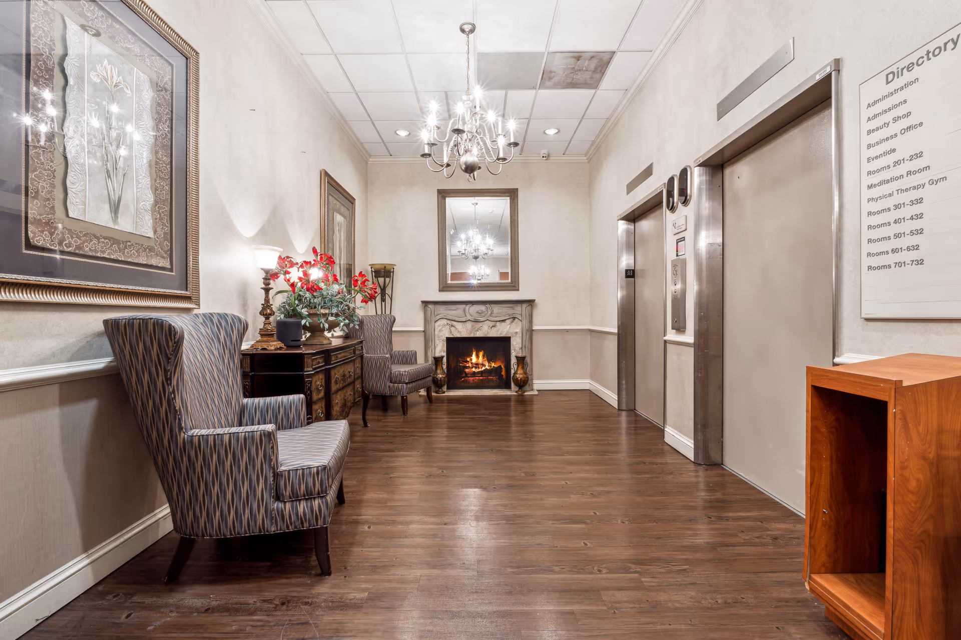 A hallway in a nursing and rehab center featuring two elevators on the right side, a wooden floor, a lit fireplace at the end with a mirror above it, two patterned armchairs and a wooden side table with a lamp and a plant on the left side, and a directory sign on the wall near the elevators.