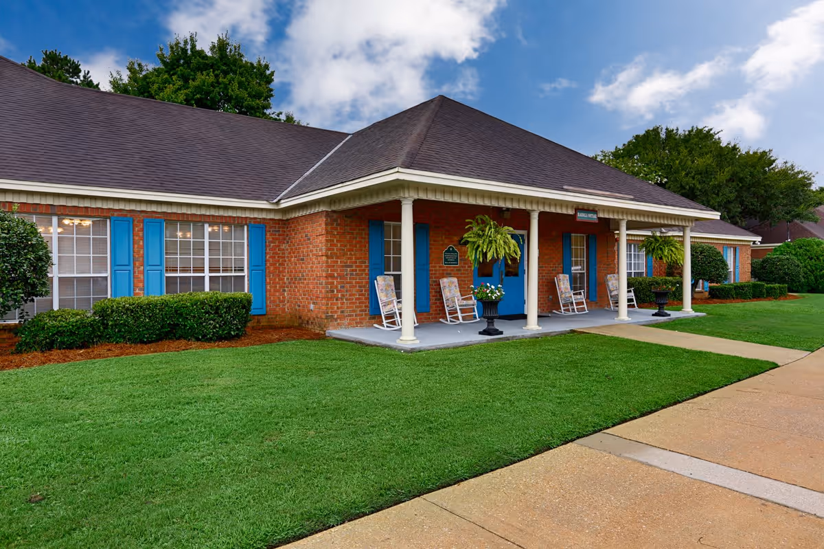 Exterior view of a single-story brick building with blue shutters and a covered porch featuring white columns and rocking chairs. The building is surrounded by neatly trimmed bushes and a well-maintained green lawn under a partly cloudy sky.