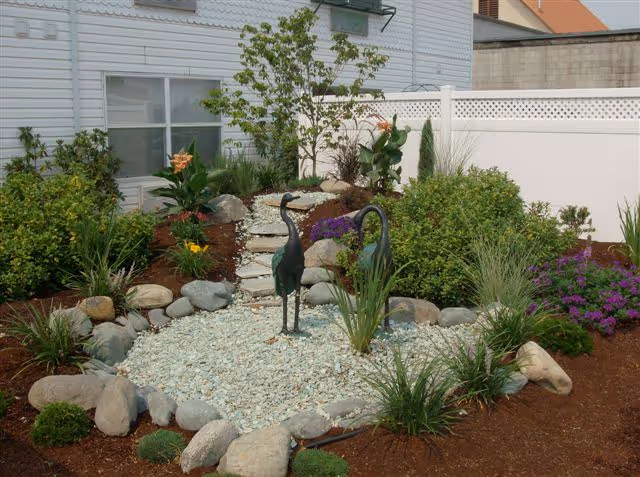 A landscaped garden area with various green plants, shrubs, and flowers surrounding a small rock bed. Two metal crane sculptures stand in the center of the rock bed. The garden is bordered by large stones and is situated next to a white building with windows and a white fence.