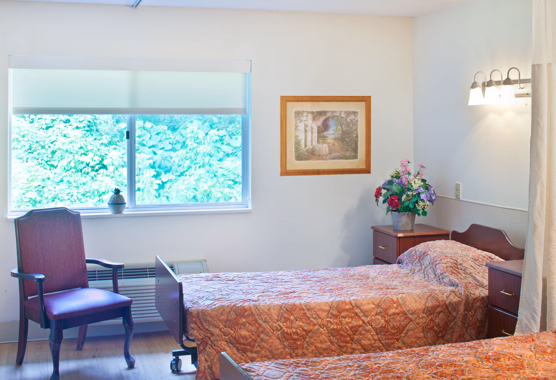 A bright room with two single beds covered in patterned bedspreads, a wooden chair with armrests near a window with a white roller shade, a framed picture on the wall, and a nightstand with a vase of colorful flowers under a wall-mounted light fixture.