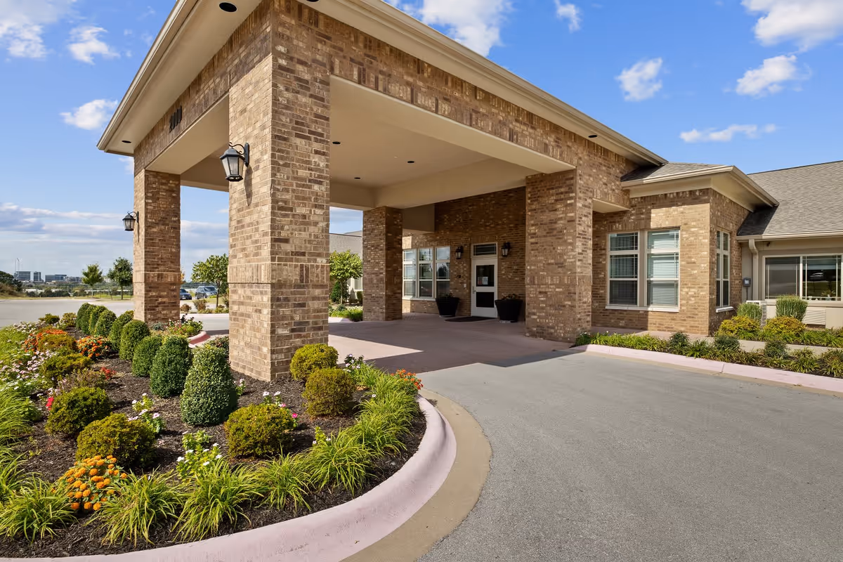 Entrance of a brick building with a covered driveway, landscaped with neatly trimmed bushes and flowers under a blue sky with some clouds.