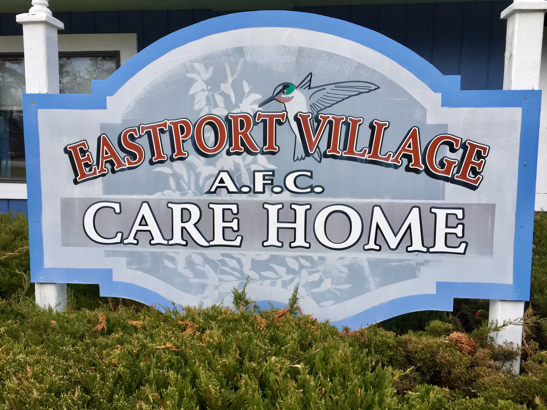 A large outdoor sign for Eastport Village A.F.C. Care Home with a blue border and a hummingbird illustration, surrounded by green bushes and part of a blue building in the background.