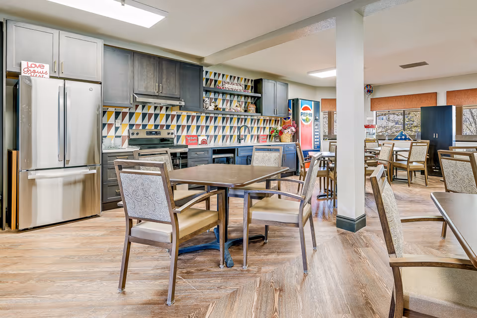 Communal dining area and kitchenette with tables and chairs, stainless steel refrigerator, colorful tiled backsplash, and a Pepsi vending machine.