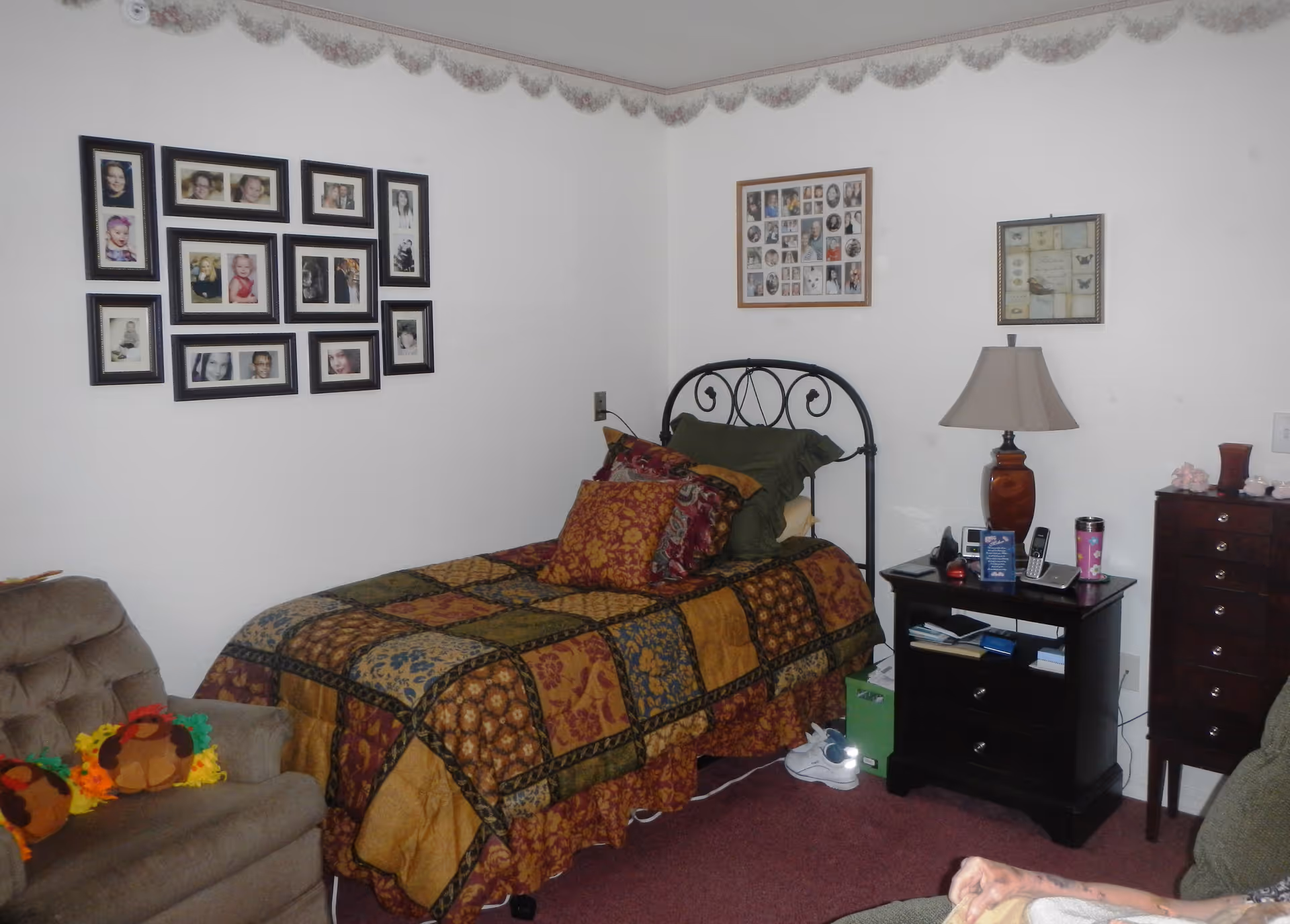 A cozy bedroom with a single bed covered in a patchwork quilt with multiple pillows. The walls are decorated with several framed photo collages. There is a nightstand with a lamp, phone, and other small items next to the bed. A cushioned armchair with colorful stuffed toys is positioned near the bed.