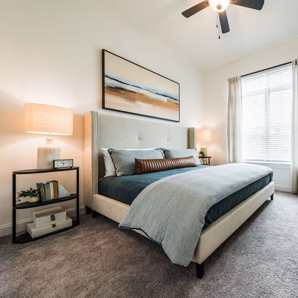A modern bedroom with a large upholstered bed featuring multiple pillows and a blue-gray comforter. There are two bedside tables with lamps, books, and decorative items. A large abstract painting hangs above the bed, and a window with blinds and light curtains lets in natural light. The room has carpeted flooring and a ceiling fan with a light.