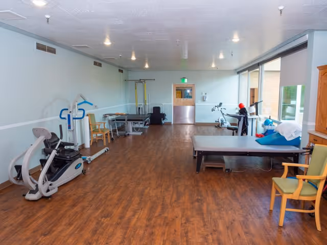 A spacious rehabilitation/exercise room with therapy equipment, treatment tables, chairs, and large windows.