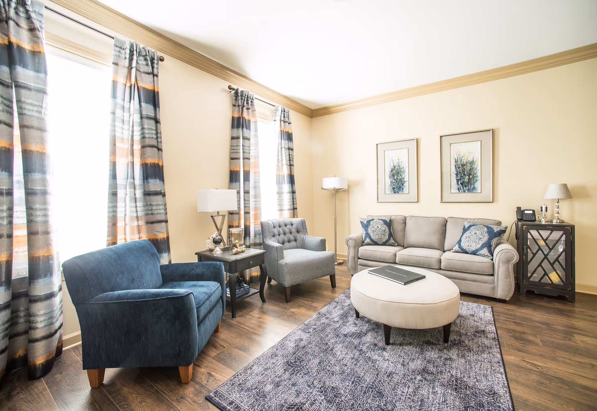 Bright living room with a sofa, two armchairs, a round ottoman, side tables and lamps, framed artwork, and patterned curtains.