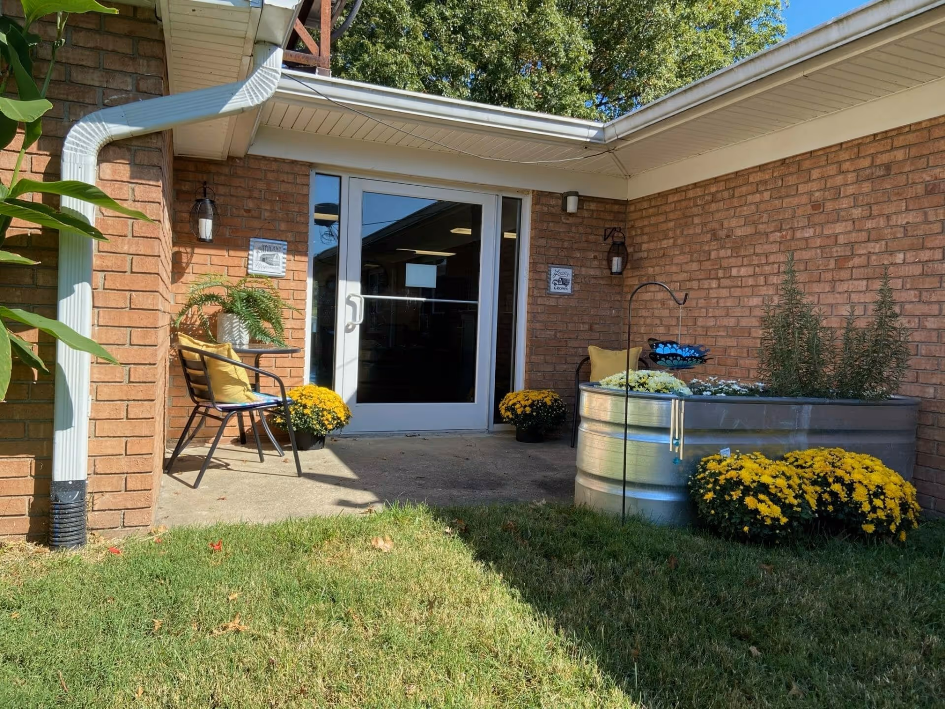 Outdoor patio area at Willowdale Village with two black metal chairs with yellow cushions, potted yellow flowers, a metal planter with various plants, and a glass door entrance set in a brick wall under a white roof overhang.