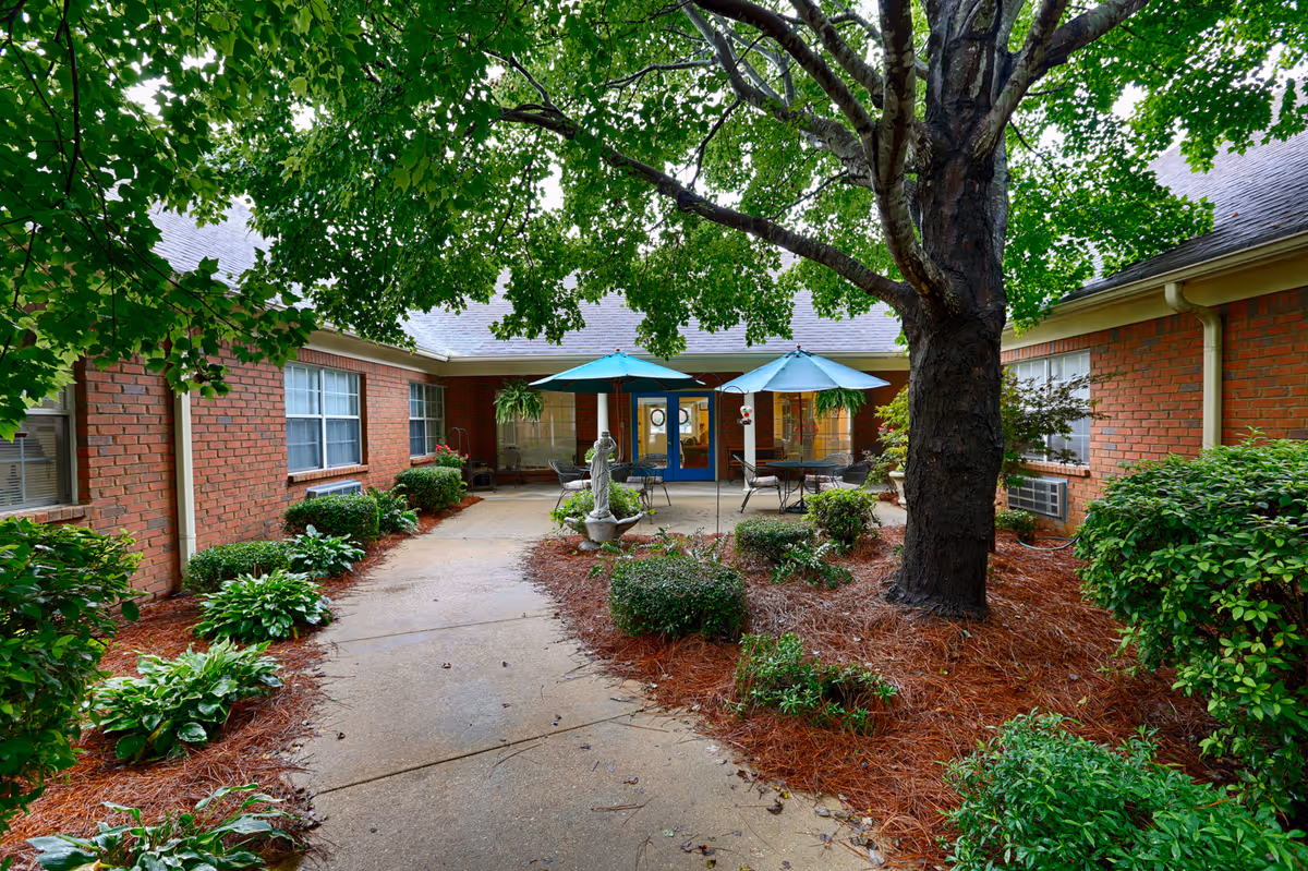 Outdoor courtyard area at Country Cottage Montgomery featuring a large tree, green shrubs, and plants along a concrete pathway. There are two patio tables with blue umbrellas and chairs near the entrance of a brick building with blue doors and windows.