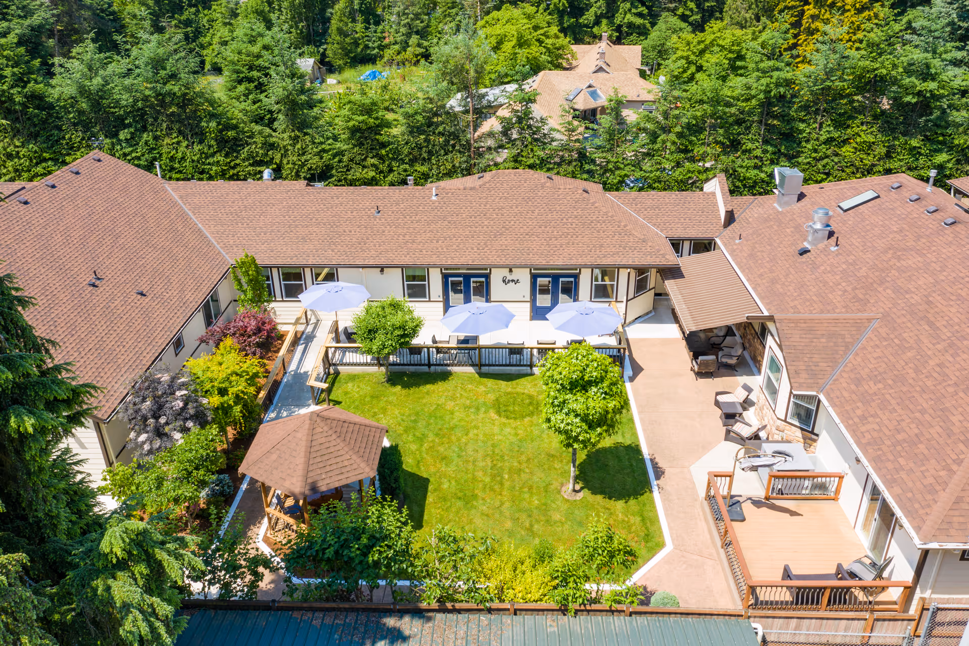 Aerial view of a senior living facility courtyard with a well-maintained green lawn, a small gazebo, several trees, and patio umbrellas providing shade over outdoor seating areas. The surrounding buildings have brown shingled roofs and beige walls, with a backdrop of dense green trees.