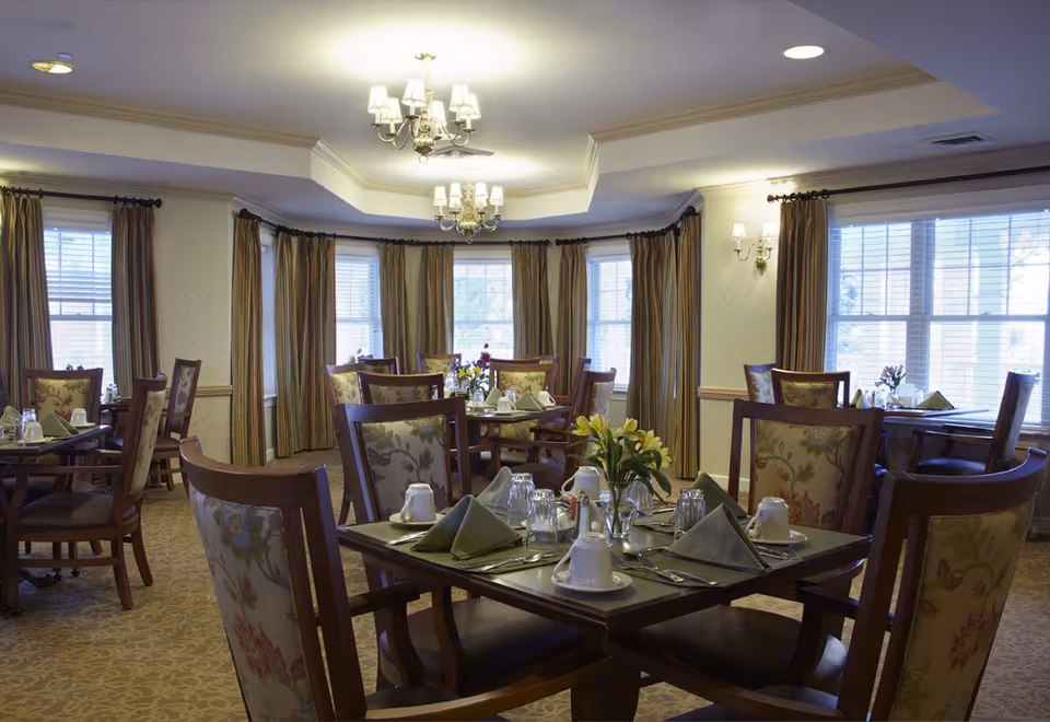 A well-lit dining room with multiple wooden tables and chairs arranged neatly. Each table is set with folded green napkins, white cups, glasses, silverware, and small flower arrangements. The room features large windows with striped curtains, beige patterned carpet, and elegant chandeliers hanging from the ceiling.