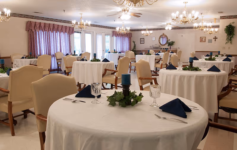 A dining room with multiple round tables covered with white tablecloths, each set with blue folded napkins, glassware, silverware, and a centerpiece featuring a blue candle surrounded by green foliage. The room has beige upholstered chairs, chandeliers hanging from the ceiling, and purple curtains covering the windows.
