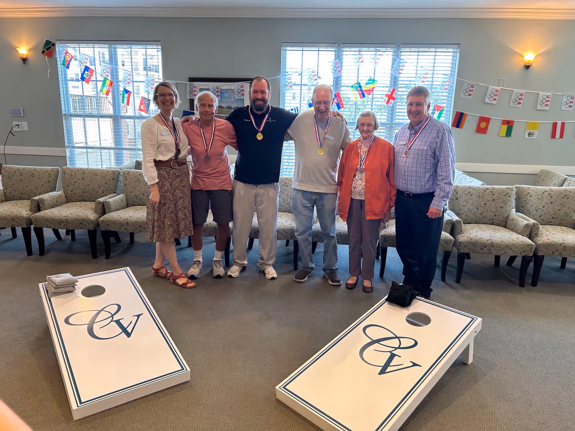 Five adults standing indoors between two cornhole boards with the letters CV on them. They are smiling and wearing medals around their necks. Behind them are several chairs and windows decorated with small international flags and USA banners.