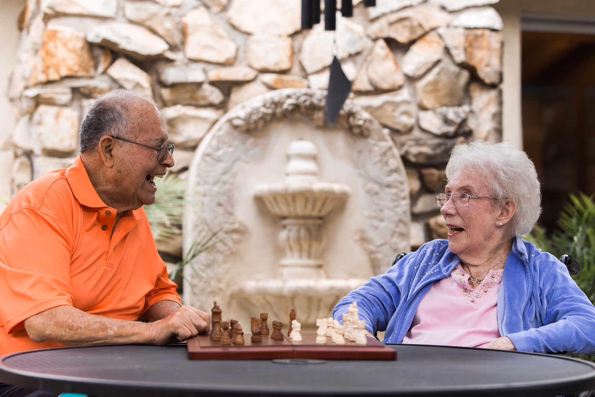 Two elderly people sitting at a round table playing chess outdoors. They are smiling and engaged in conversation. Behind them is a stone wall with a decorative water fountain.