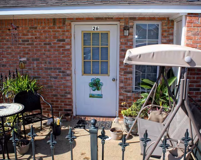 A small outdoor patio area in front of a white door numbered 26 on a red brick building. The patio features a round table with two black chairs, several potted plants, and a beige cushioned swing. There is a black metal fence in the foreground.