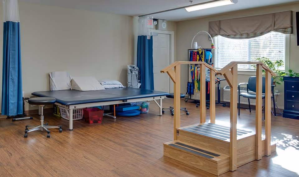 A physical therapy room with a padded treatment table, a stool on wheels, a wooden step exercise platform with handrails, and various therapy equipment near a window with blinds. The room has wood flooring and light-colored walls.