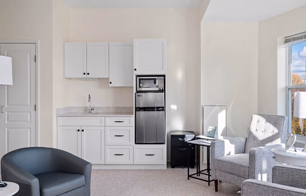 A bright and clean interior space featuring a small kitchenette with white cabinets, a sink, a microwave, and a mini refrigerator. To the right, there is a gray upholstered armchair next to a window with natural light streaming in, accompanied by a small black side table and a white round table with a lamp. On the left, there is a dark gray armchair and a closed white door.