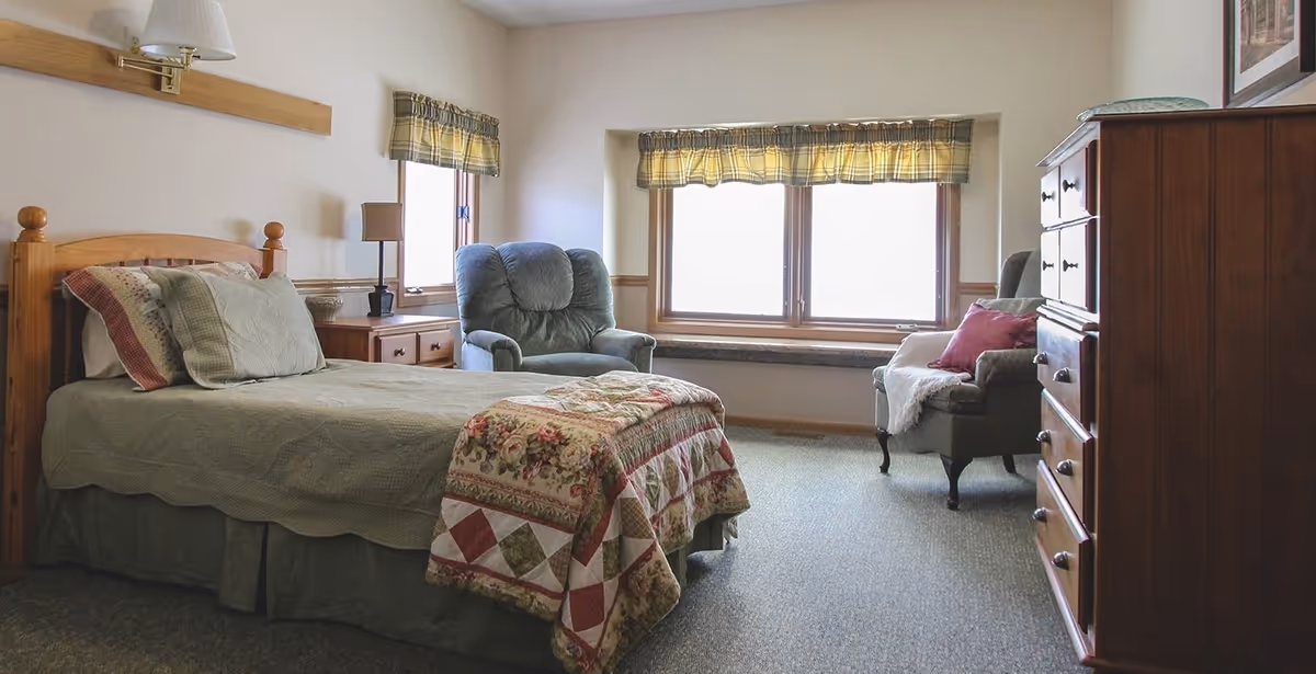 A cozy bedroom with a single bed covered in a quilted blanket and pillows, a wooden nightstand with a lamp, a comfortable armchair, a window with plaid curtains, and a wooden dresser.