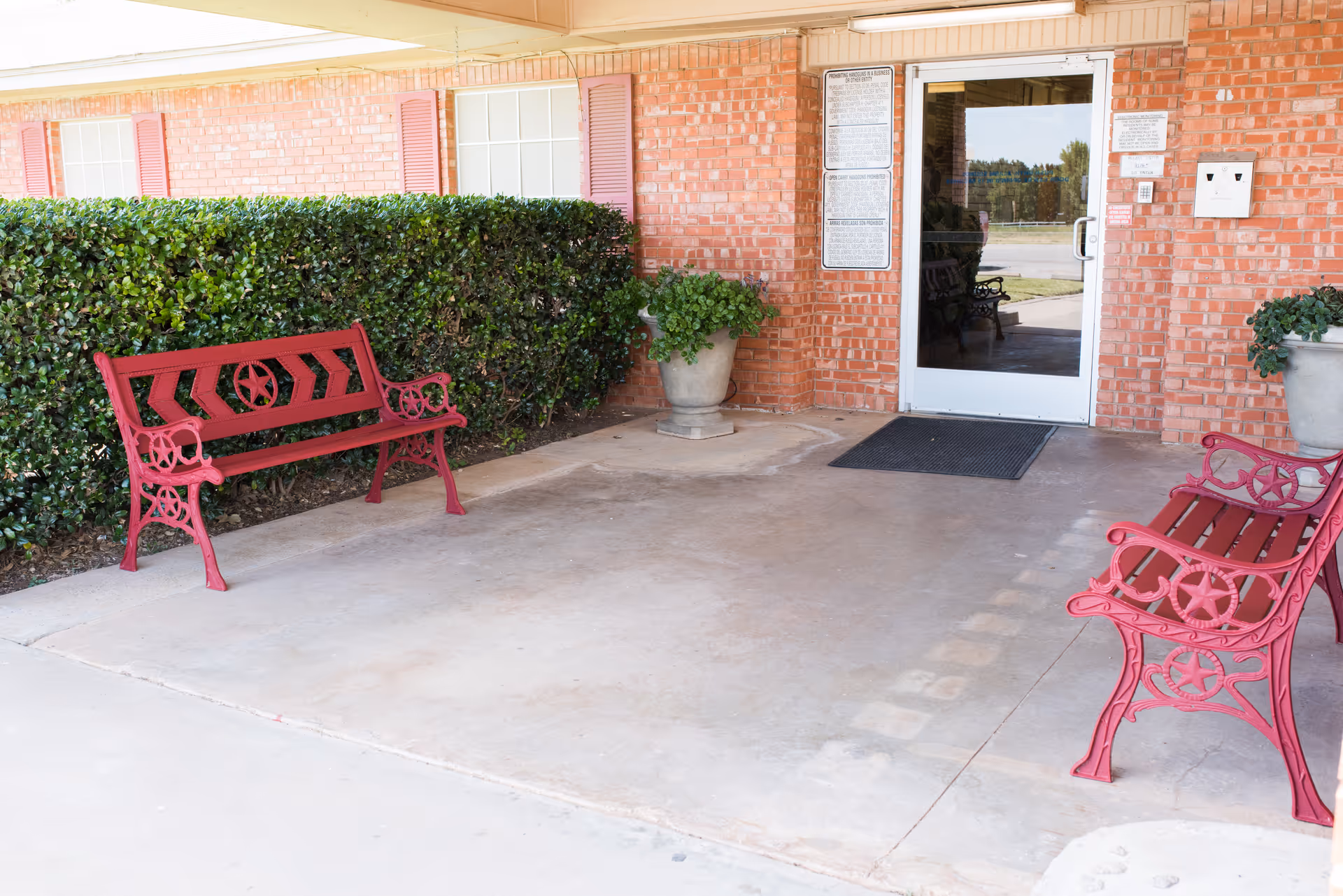 Outdoor covered patio area with two red metal benches facing each other, a hedge along the brick wall, two large potted plants, and a glass door entrance to the building.