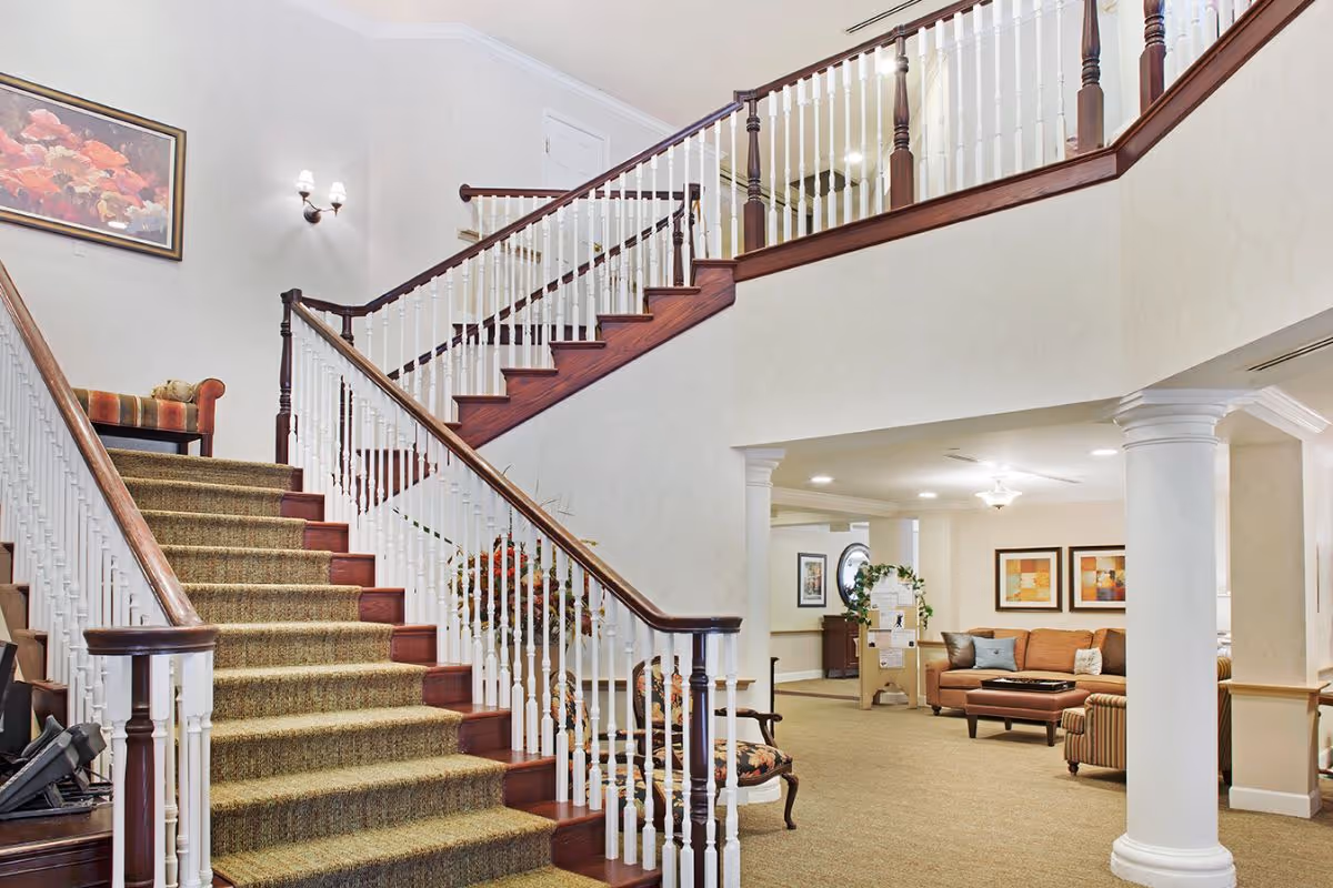 Carpeted central staircase with white balusters overlooking a furnished lounge area with couches and columns.