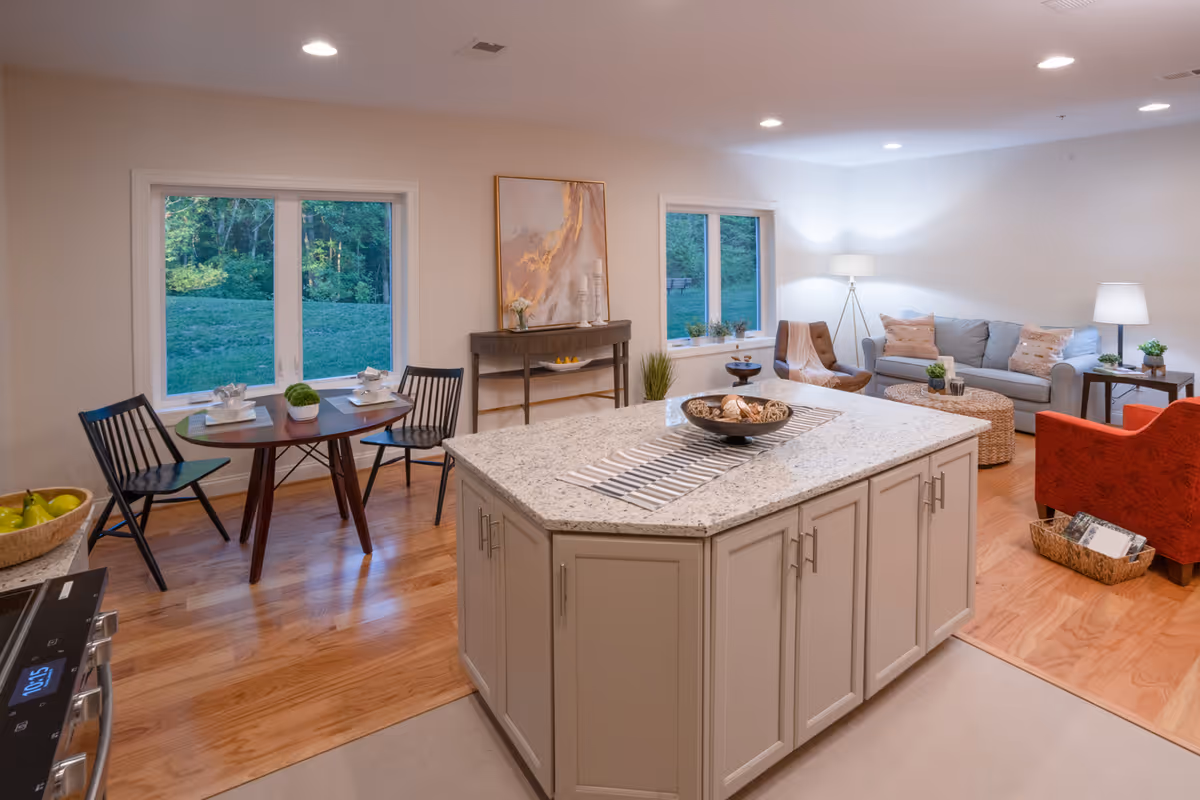 A bright and spacious open-concept living area in a retirement community featuring a kitchen island with a granite countertop in the foreground, a small dining table with two chairs near two large windows showing greenery outside, and a cozy living room area with a gray sofa, an orange armchair, a brown chair, two side tables with lamps, and a floor lamp. The room has wooden flooring and recessed ceiling lights.