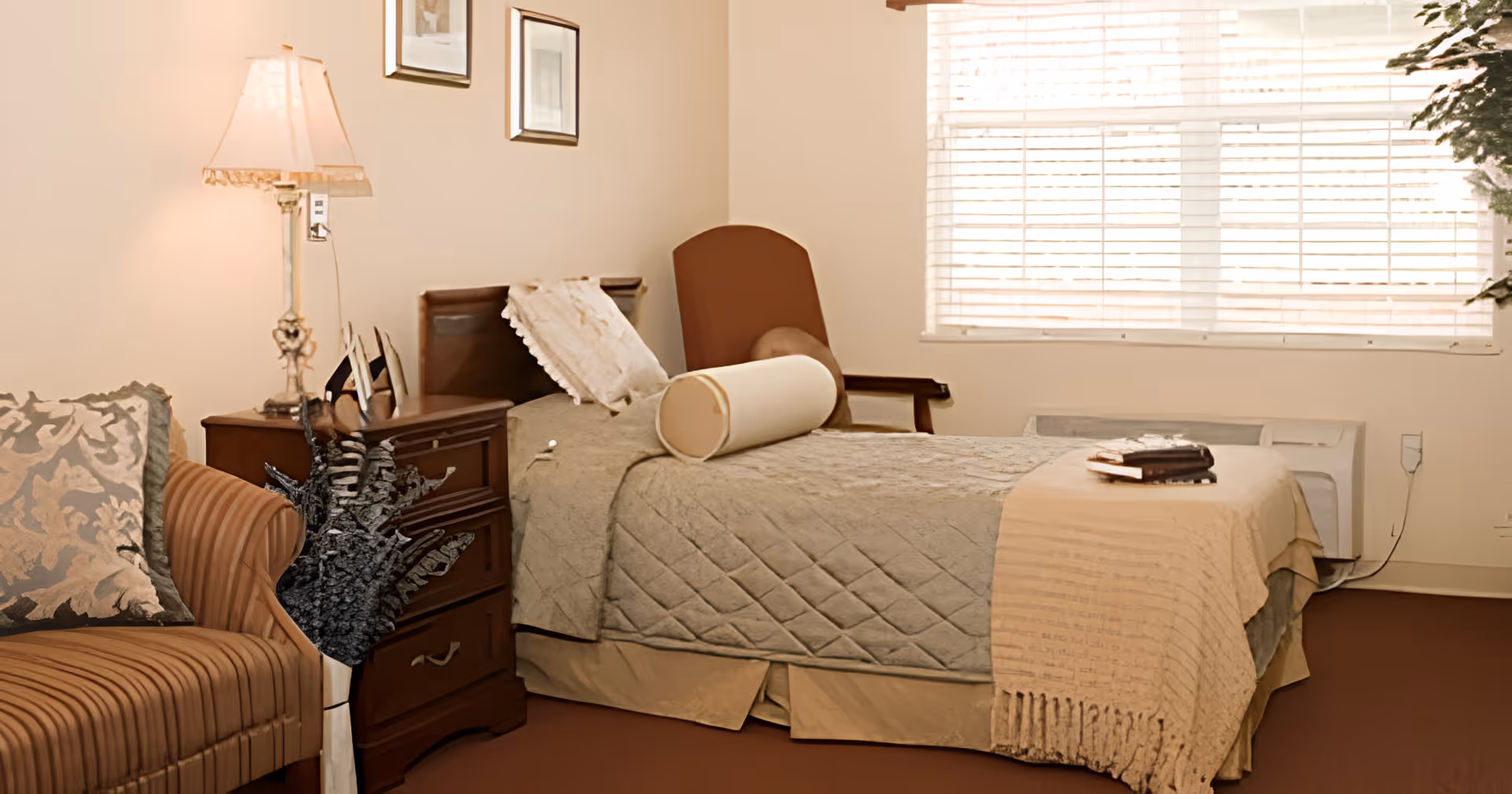 A cozy bedroom with a single bed covered in a quilted bedspread and a cylindrical pillow. Next to the bed is a wooden nightstand with a decorative lamp and some framed pictures on the wall above. There is a cushioned armchair with patterned pillows on the left side and a window with blinds letting in natural light on the right side.