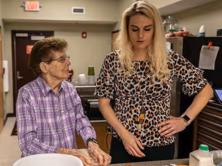 An elderly woman wearing a purple plaid shirt and a younger woman with long blonde hair wearing a leopard print blouse are standing together in a kitchen area. The elderly woman is looking at the younger woman, who is focused on something on the counter in front of her. Various kitchen items and appliances are visible in the background.