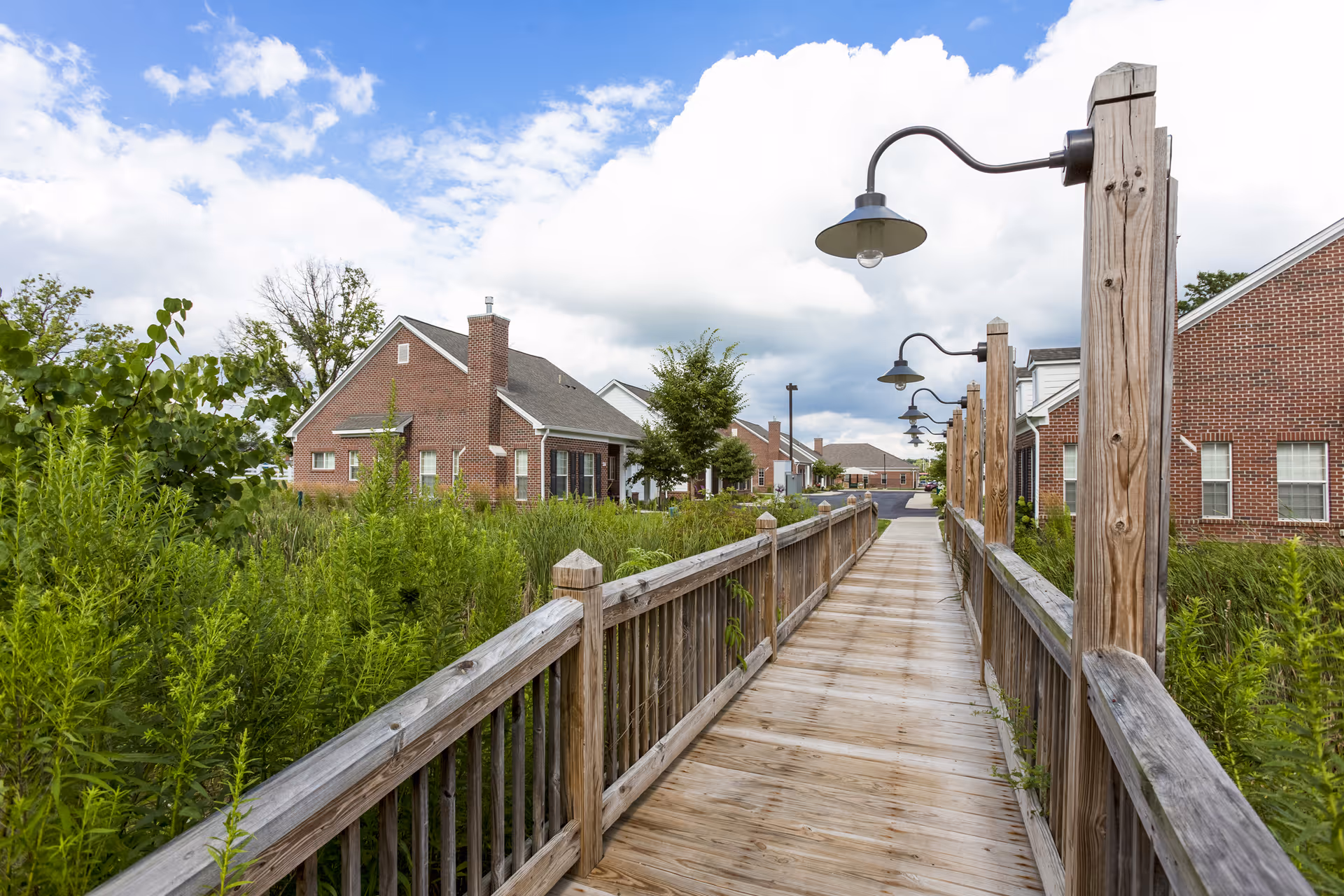 A wooden boardwalk with railings and vintage-style lamp posts on the right side, leading through lush green vegetation towards brick buildings under a partly cloudy blue sky.