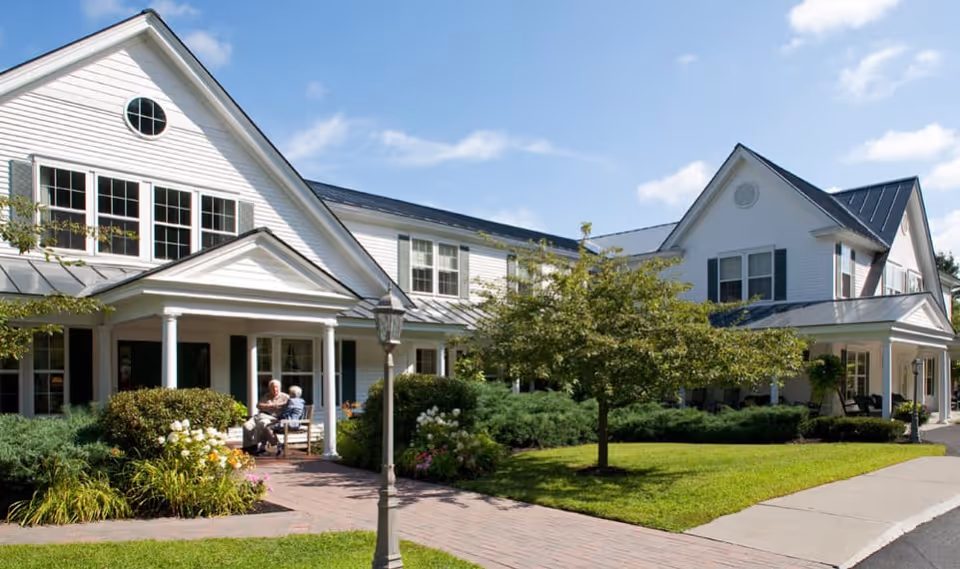 Exterior view of a white two-story senior living facility with green shutters and a covered porch. Two elderly people are sitting and conversing on chairs on the porch. The surrounding area features well-maintained landscaping with bushes, flowers, a tree, and a brick walkway leading to the entrance.
