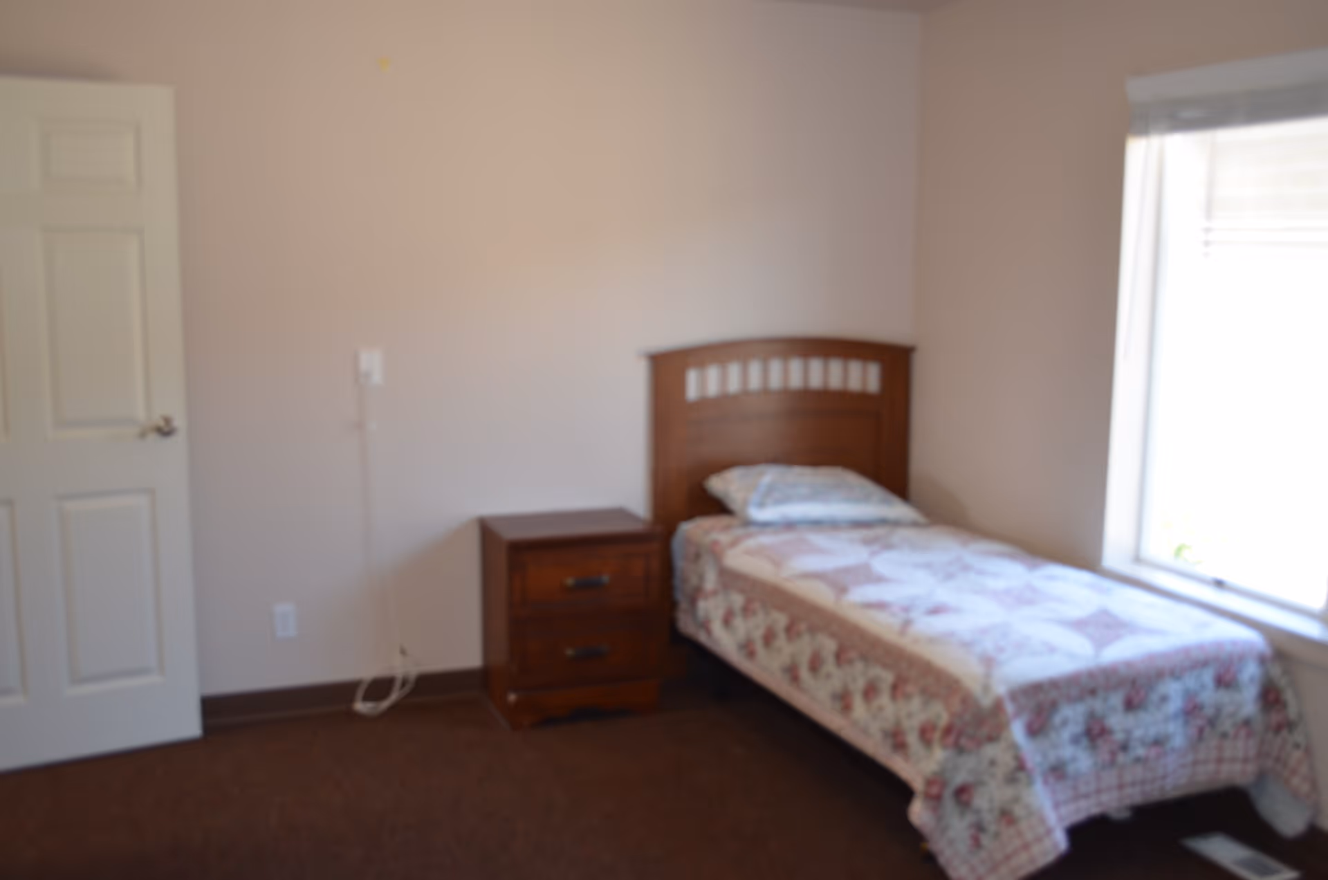 A simple bedroom with a single wooden bed covered with a floral quilt and a pillow. Next to the bed is a wooden nightstand with two drawers. There is a window with blinds on the right side letting in natural light, and a closed white door on the left side of the room. The walls are light-colored and the floor is carpeted.