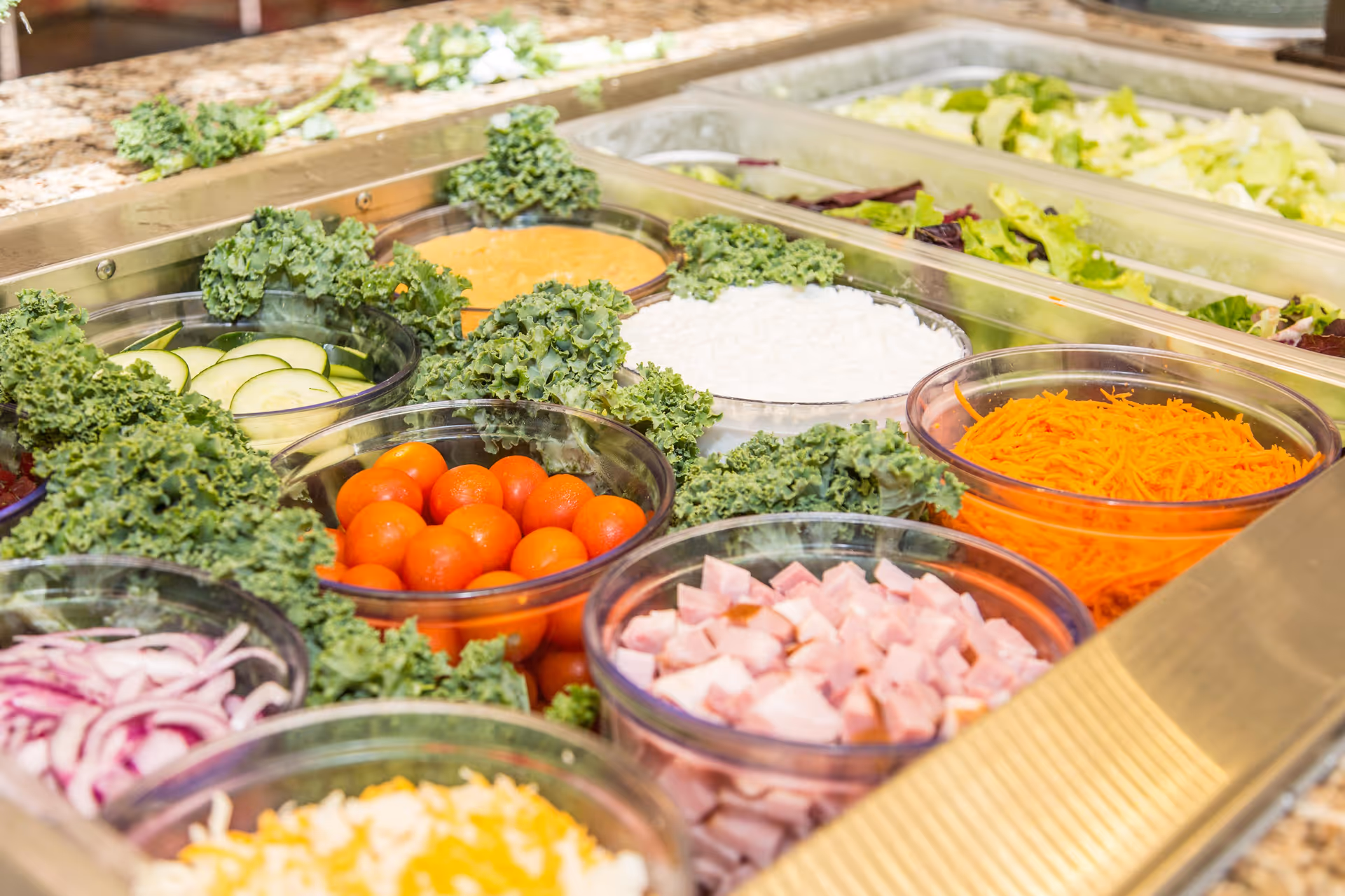 Self-serve salad bar with bowls of cherry tomatoes, shredded carrots, diced ham, sliced cucumbers, onions, cheese and leafy greens.