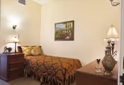 Small furnished bedroom with a single bed, nightstand, dresser, lamps, and framed wall art.